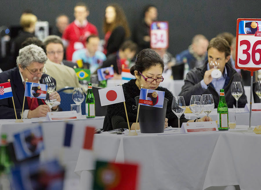 Picture of judges from several countries tasting wine. There is a flag from the judge's country on his table.
