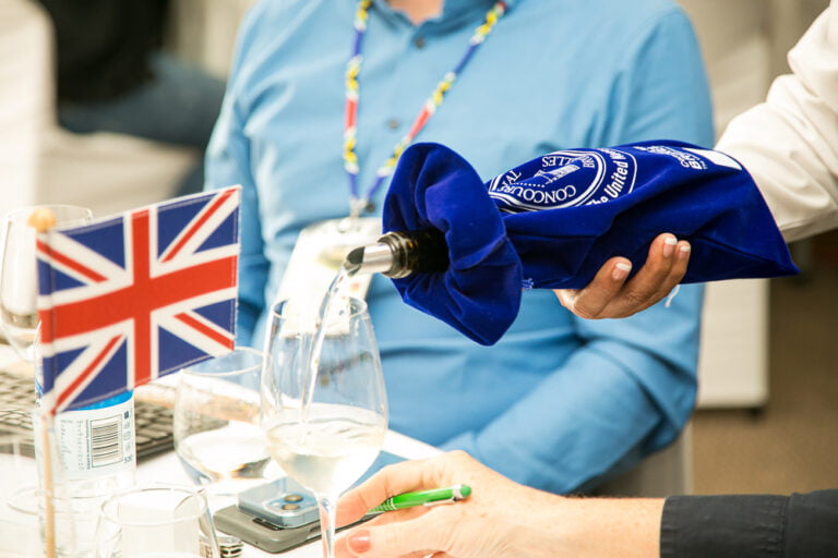 Jemand serviert Weißwein in einem Weinglas. Die Flasche ist in einer blauen CMB-markierten Stofftasche versteckt. Auf dem Tisch steht eine England-Flagge.